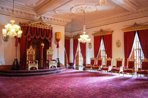 A grand room featuring ornate throne chairs, elegant chandeliers, and rich red drapes, creating a regal atmosphere.