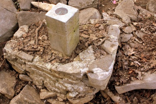 A weathered stone structure with a concrete pillar on top, surrounded by leaves and rocks in a natural setting.