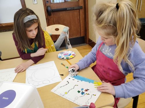 Two children engaged in an art activity, one painting while the other observes, with colorful supplies on the table.