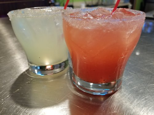 Two colorful cocktails in glasses with ice, one light green and one red, both with salt rims and straws.