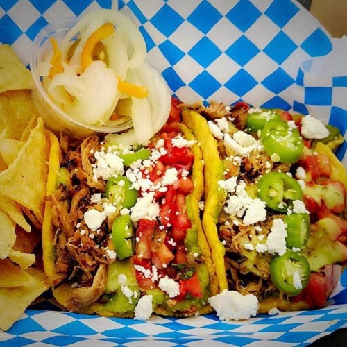 A colorful plate featuring three tacos topped with tomatoes, jalapeños, and cheese, served with tortilla chips and pickled vegetables.