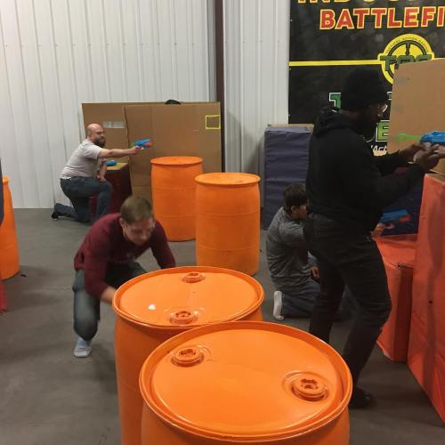 A group of people playing a game in an indoor arena, using orange barrels and cardboard for cover while holding toy guns.