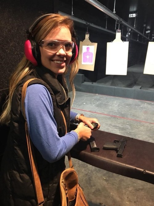A woman wearing safety glasses and ear protection smiles while handling firearms at a shooting range.