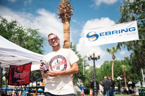 A smiling man in sunglasses holds a soda bottle at a festival under a blue sky with palm trees and a banner.
