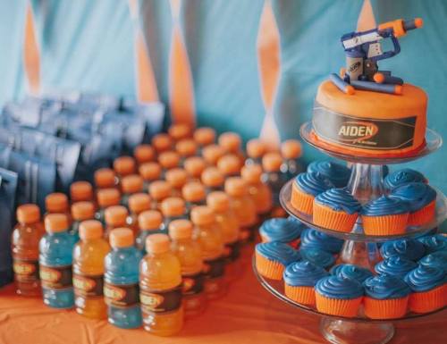 A colorful birthday setup featuring a cake with a toy gun, blue cupcakes, and orange drinks on a table.