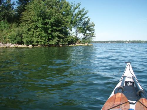 A kayak on calm water, with lush green trees lining the shore under a clear blue sky.