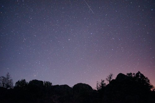 A starry night sky filled with twinkling stars above dark silhouettes of rocky terrain and trees.