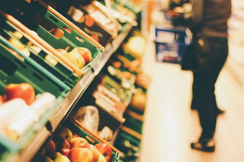 A grocery store aisle with green crates of apples and a blurred figure in the background holding a shopping basket.