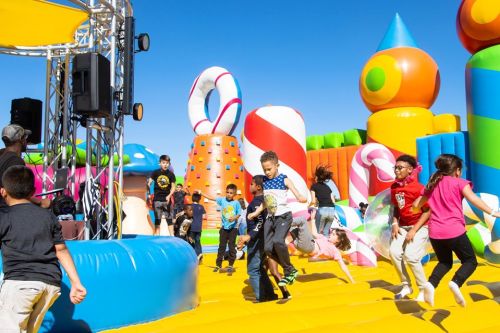 Children playing and jumping on a colorful inflatable playground with candy-themed structures under a clear blue sky.