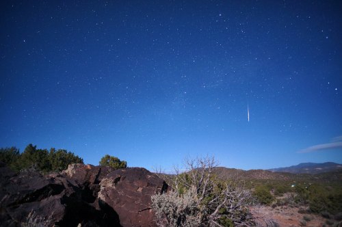 A clear night sky filled with stars, featuring a shooting star above rocky terrain and distant mountains.