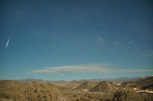 A starry night sky over mountains, featuring a bright meteor streaking across the scene.