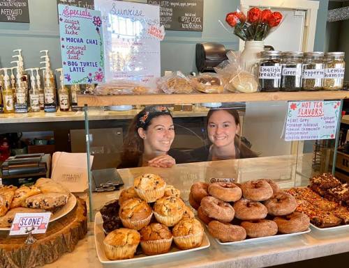 Two smiling baristas stand behind a display case filled with muffins, donuts, and baked goods in a cozy café.