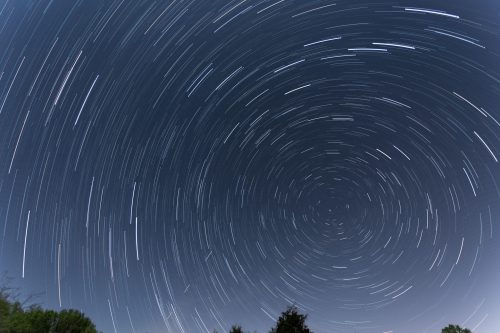 Star trails create a circular pattern in the night sky, with dark silhouettes of trees at the bottom.