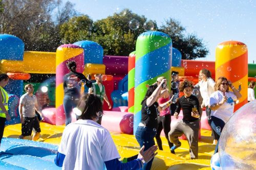 A lively scene of people playing on colorful inflatable structures, with bubbles floating in the air on a sunny day.