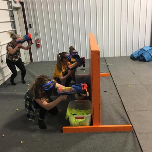 Four people wearing protective goggles aim toy blasters over an orange barrier in a playful indoor setting.