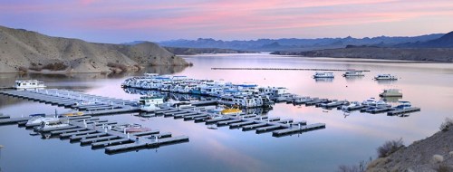 A serene marina at sunset, with boats docked and mountains in the background reflecting on calm waters.