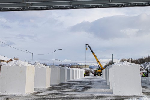 A crane lifts a block of snow in a snowy area with large white snow blocks under a bridge and mountains in the background.