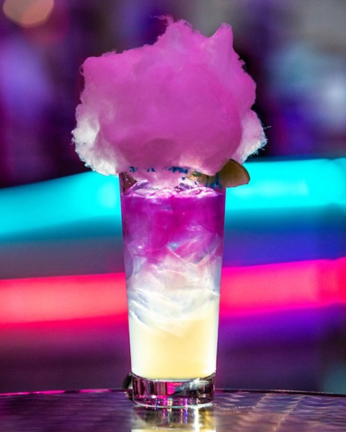 A colorful cocktail topped with fluffy pink cotton candy, set against a vibrant, illuminated background.