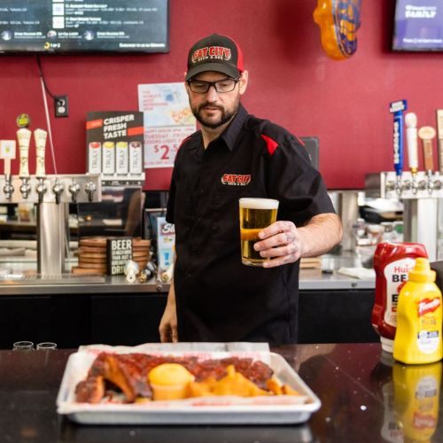 A man in a black shirt holds a beer while standing behind a bar with a platter of ribs and sides in front of him.