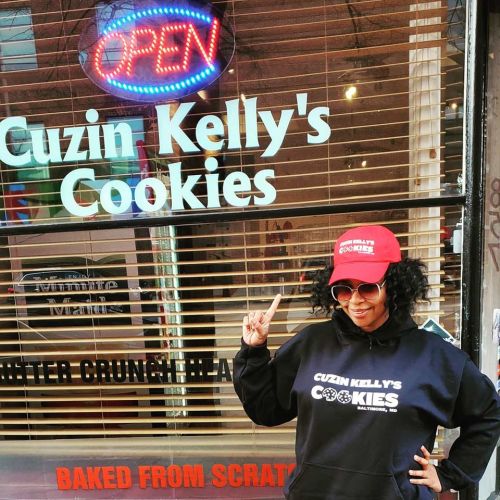 A person in a red hat and black hoodie poses in front of "Cuzin Kelly's Cookies" shop with an open sign.