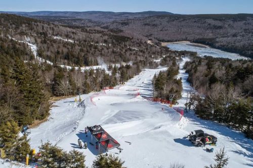 A snowy landscape with ski slopes, trees, and a distant lake under a clear blue sky. Equipment is visible on the slopes.