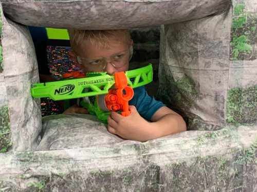 A young boy aims a Nerf blaster from a play structure, wearing safety goggles and focused on his target.