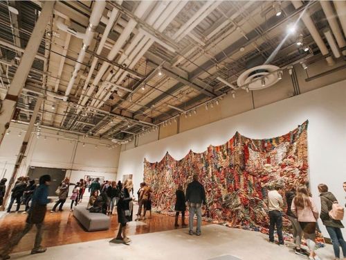 A vibrant textile art installation on a gallery wall, with visitors admiring the piece in a spacious, modern art space.