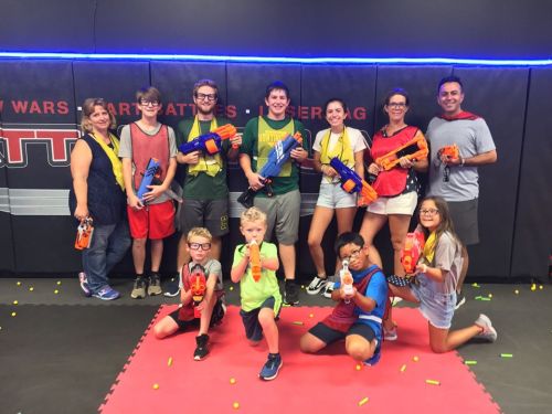 A group of children and adults pose with toy blasters in a colorful indoor play area, surrounded by foam darts.