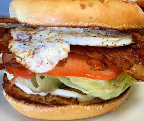 A close-up of a sandwich with bacon, lettuce, tomato, a fried egg, and mayonnaise on a toasted sesame seed bun.