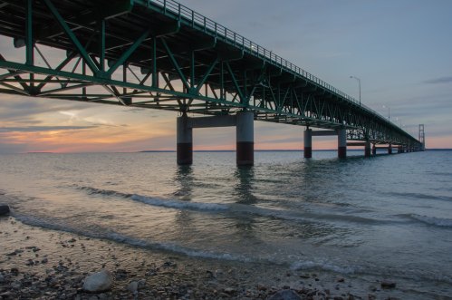 A green bridge spans over calm waters at sunset, with gentle waves lapping at the rocky shore below.