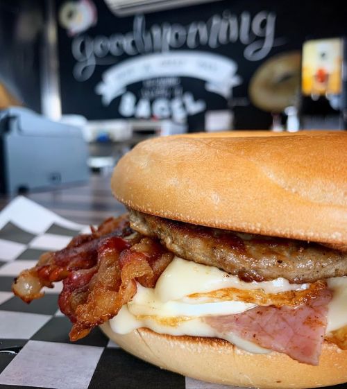 A close-up of a breakfast sandwich with sausage, bacon, and egg on a bagel, set against a chalkboard background.