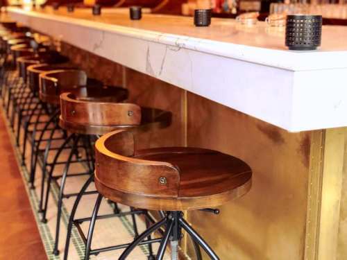 A row of wooden bar stools with metal bases lined up against a marble-topped bar.