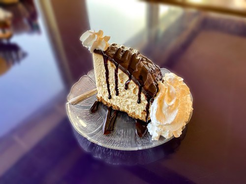 A slice of cheesecake topped with chocolate drizzle and whipped cream on a clear plate.