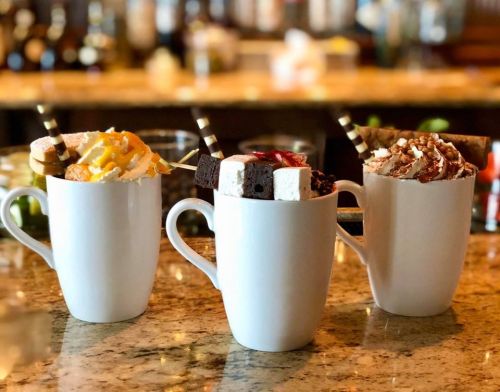 Three decorative mugs filled with colorful desserts, topped with whipped cream, chocolate, and various garnishes.