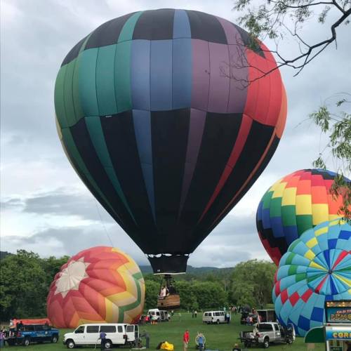 Colorful hot air balloons inflate in a grassy field, with people and vehicles nearby under a cloudy sky.