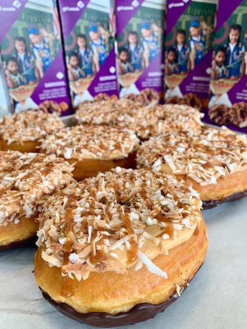 A close-up of donuts topped with caramel, coconut, and chocolate, with boxes of cookies in the background.