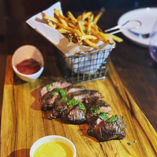A wooden platter with sliced steak topped with green sauce, accompanied by a basket of fries and dipping sauces.