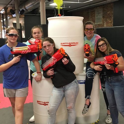 Five girls pose with toy blasters in front of stacked barrels, wearing protective eyewear in a playful indoor setting.
