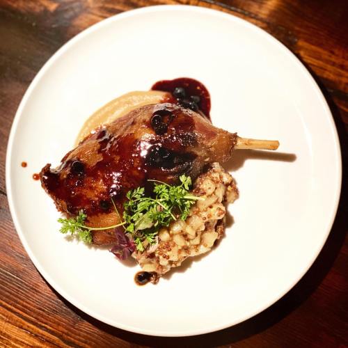 A plated dish featuring duck leg with a glossy sauce, served with grain and a side of puree, garnished with herbs.