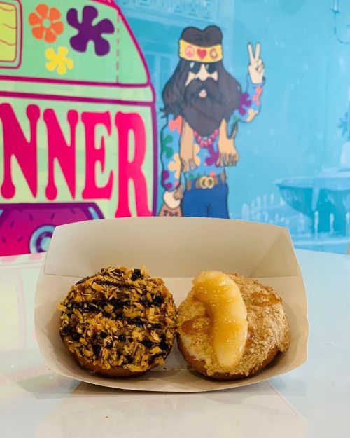 Two donuts in a white box: one topped with coconut and chocolate, the other with banana and cinnamon, against a colorful backdrop.
