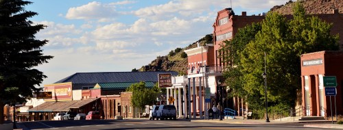 A historic street lined with brick buildings, trees, and a clear blue sky with scattered clouds.