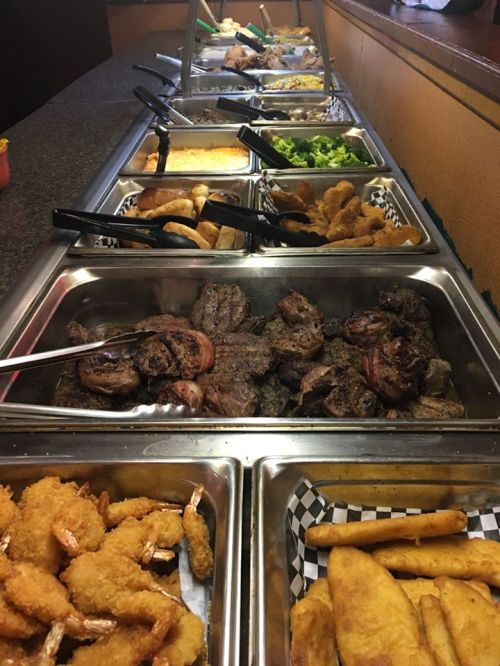 A buffet spread featuring various meats, shrimp, sides, and salads in metal trays with serving utensils.