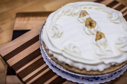 A creamy banana cream pie topped with whipped cream and banana slices, placed on a wooden cutting board.