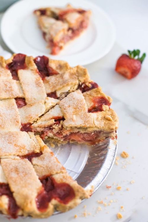 A freshly baked strawberry pie with a lattice crust, partially sliced, on a marble surface with a plate of pie in the background.