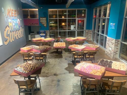 A person stands in a donut shop surrounded by large boxes of colorful donuts on tables. Brightly lit and inviting atmosphere.