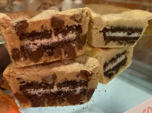 Close-up of layered dessert bars with chocolate chip cookie dough and Oreo cookies inside, displayed in a glass case.