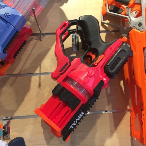 A colorful toy blaster with a red barrel and black handle, displayed on a shelf among other toy guns.