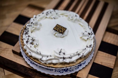 A round cake with white frosting, decorative swirls, and a chocolate square on top, placed on a wooden surface.