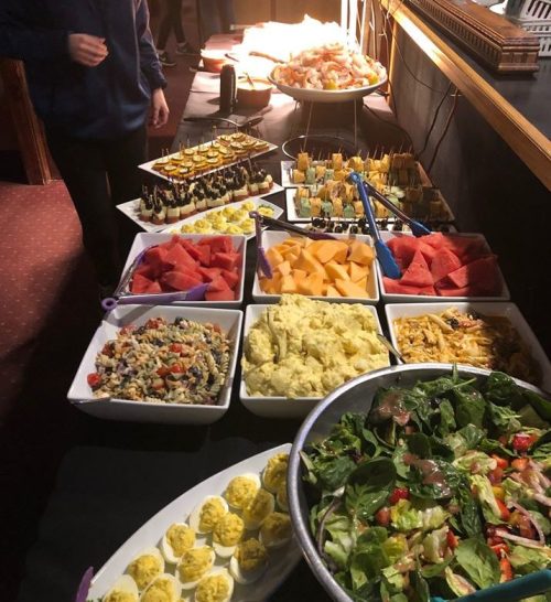A buffet table with various dishes, including salads, fruits, and appetizers, with a person serving food in the background.