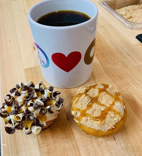 A cup of black coffee next to two donuts: one topped with chocolate curls and caramel, the other with a crumb topping.